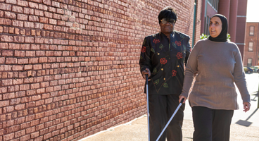 Two diverse blind woman walk along a sidewalk using their long white canes.