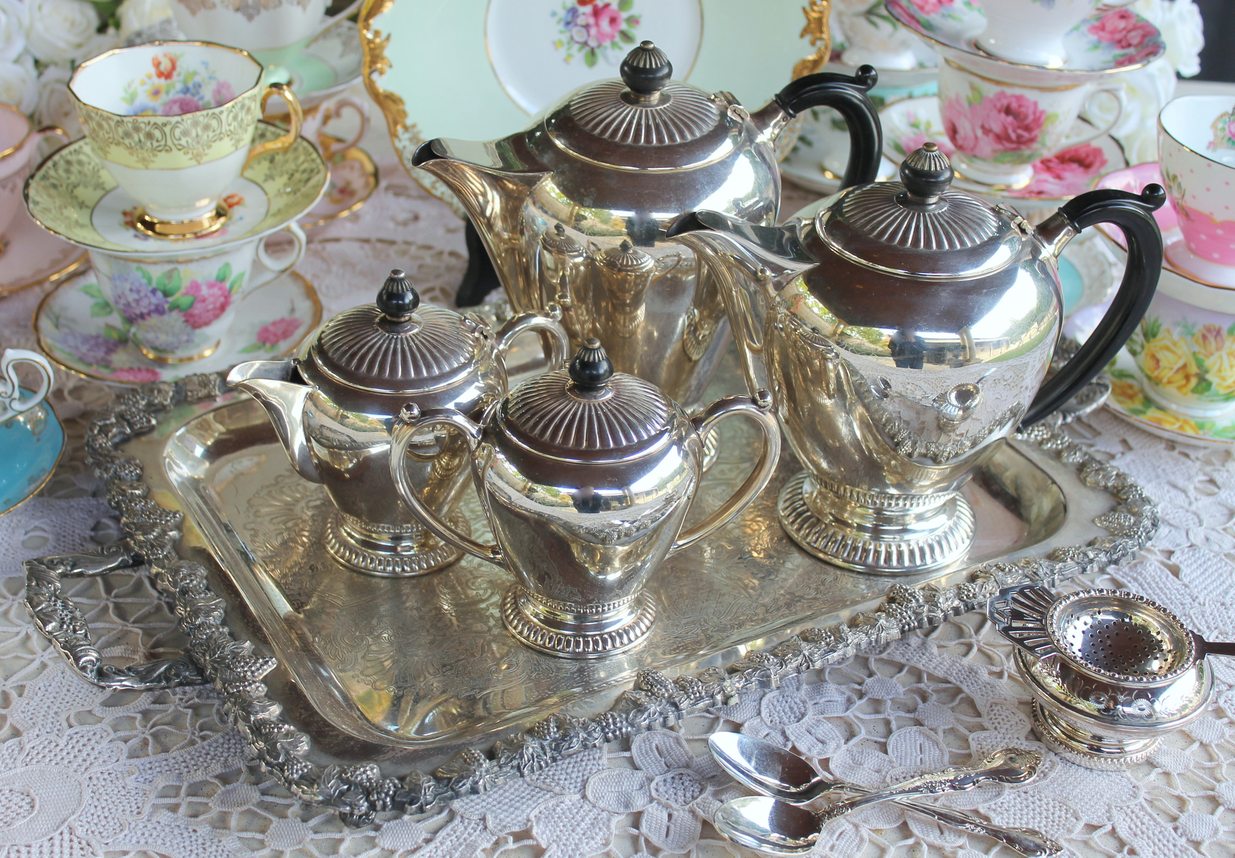 Silver tea service on a lace tablecloth surrounded by cups on saucers
